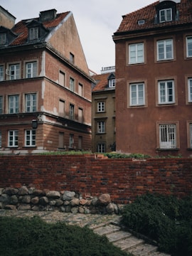 Historic buildings with brown and red facades, featuring white windows, are situated behind a red brick wall. The roofs are covered with reddish-brown tiles. In front of the wall, there is a small patch of greenery and a stone pathway.
