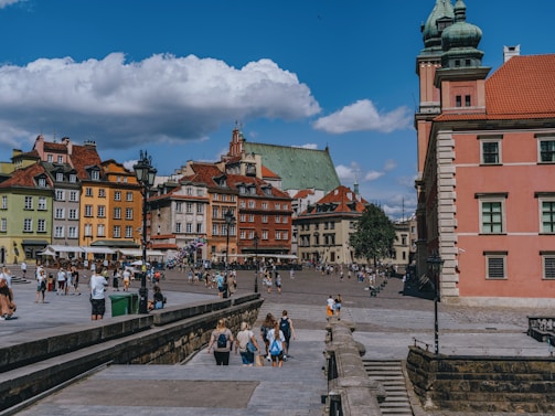 A bustling town square featuring colorful historic buildings with red, green, and yellow facades. Crowds of people, including tourists and locals, mingle and explore the area. The sky is blue with scattered clouds, adding to the lively yet relaxed atmosphere. A large church with a green roof is visible in the background.