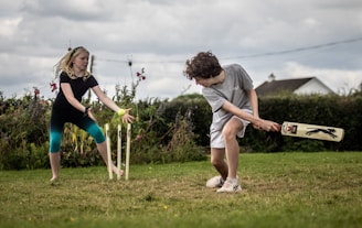 A boy and a girl are playing cricket on a grassy field. The boy is batting, wearing a grey t-shirt and white shorts, and is holding a cricket bat. The girl, dressed in a black t-shirt and blue leggings, is fielding near the stumps. There are wildflowers and a hedge in the background, with a house visible in the distance.