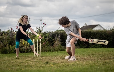 A boy and a girl are playing cricket on a grassy field. The boy is batting, wearing a grey t-shirt and white shorts, and is holding a cricket bat. The girl, dressed in a black t-shirt and blue leggings, is fielding near the stumps. There are wildflowers and a hedge in the background, with a house visible in the distance.