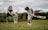A boy and a girl are playing cricket on a grassy field. The boy is batting, wearing a grey t-shirt and white shorts, and is holding a cricket bat. The girl, dressed in a black t-shirt and blue leggings, is fielding near the stumps. There are wildflowers and a hedge in the background, with a house visible in the distance.