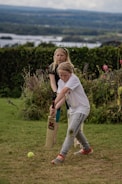 Two children are playing cricket on a grassy area with one holding a cricket bat and the other standing behind. The background features lush greenery and a distant view of a lake under a cloudy sky.