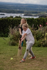 Two children are playing cricket on a grassy area with one holding a cricket bat and the other standing behind. The background features lush greenery and a distant view of a lake under a cloudy sky.