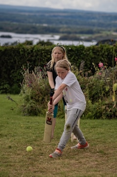 Two children are playing cricket on a grassy area with one holding a cricket bat and the other standing behind. The background features lush greenery and a distant view of a lake under a cloudy sky.