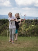 Two young girls are playing cricket on a grassy field. One girl is swinging a cricket bat at a ball, while the other watches. The background features a hedge and some flowers, with a cloudy sky above.