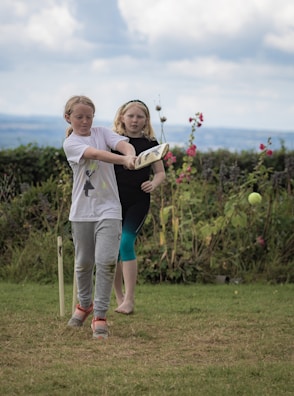 Two young girls are playing cricket on a grassy field. One girl is swinging a cricket bat at a ball, while the other watches. The background features a hedge and some flowers, with a cloudy sky above.