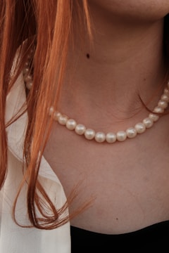 A close-up of a necklace with white pearls resting on a person's neck. The person has long, reddish hair partially covering the pearls. The clothing is partially visible, consisting of a light-colored garment on one side and a black one on the other.