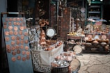 A market stall displays a variety of handcrafted metal items including trays, pots, dishes, cups, and decorative items. The items, predominantly copper-toned, are neatly arranged on tables and shelves. Vibrant textiles and patterned tablecloths serve as the backdrop for the display.
