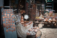 A market stall displays a variety of handcrafted metal items including trays, pots, dishes, cups, and decorative items. The items, predominantly copper-toned, are neatly arranged on tables and shelves. Vibrant textiles and patterned tablecloths serve as the backdrop for the display.