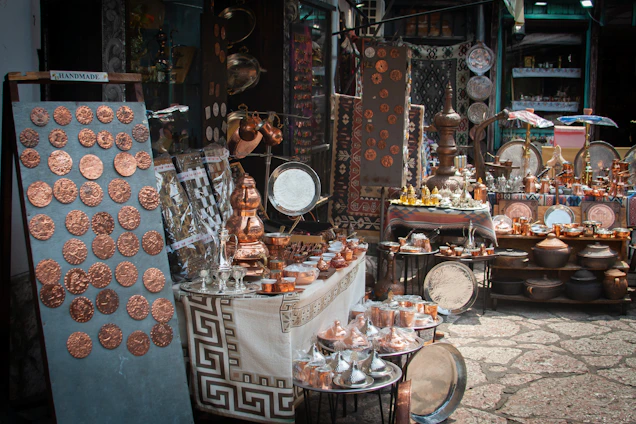 A market stall displays a variety of handcrafted metal items including trays, pots, dishes, cups, and decorative items. The items, predominantly copper-toned, are neatly arranged on tables and shelves. Vibrant textiles and patterned tablecloths serve as the backdrop for the display.