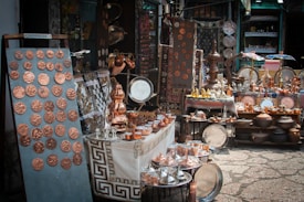 A market stall displays a variety of handcrafted metal items including trays, pots, dishes, cups, and decorative items. The items, predominantly copper-toned, are neatly arranged on tables and shelves. Vibrant textiles and patterned tablecloths serve as the backdrop for the display.
