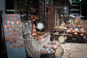 A market stall displays a variety of handcrafted metal items including trays, pots, dishes, cups, and decorative items. The items, predominantly copper-toned, are neatly arranged on tables and shelves. Vibrant textiles and patterned tablecloths serve as the backdrop for the display.