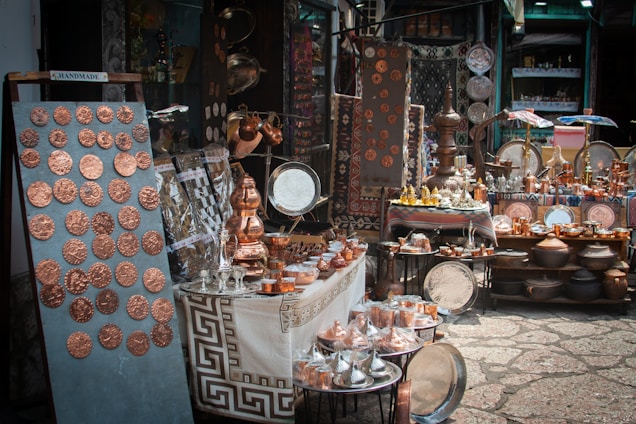 A market stall displays a variety of handcrafted metal items including trays, pots, dishes, cups, and decorative items. The items, predominantly copper-toned, are neatly arranged on tables and shelves. Vibrant textiles and patterned tablecloths serve as the backdrop for the display.