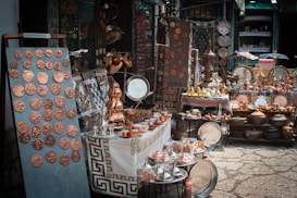 A market stall displays a variety of handcrafted metal items including trays, pots, dishes, cups, and decorative items. The items, predominantly copper-toned, are neatly arranged on tables and shelves. Vibrant textiles and patterned tablecloths serve as the backdrop for the display.