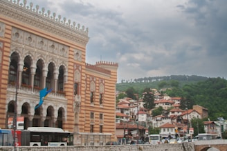 A large, intricately designed building with a mix of Moorish and Gothic architectural styles stands prominently. The facade features arched windows and detailed stonework. A blue and yellow flag is visible. In the background, a collection of smaller, red-roofed houses is nestled on a greenery-covered hillside under an overcast sky. Vehicles, including buses, are moving in front of the building.