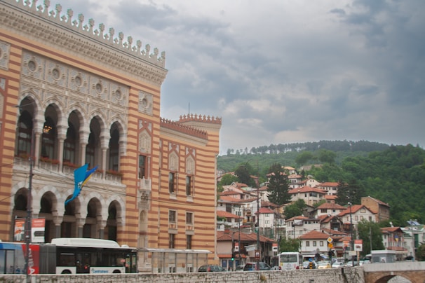 A large, intricately designed building with a mix of Moorish and Gothic architectural styles stands prominently. The facade features arched windows and detailed stonework. A blue and yellow flag is visible. In the background, a collection of smaller, red-roofed houses is nestled on a greenery-covered hillside under an overcast sky. Vehicles, including buses, are moving in front of the building.