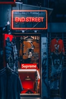 A dimly lit store entrance with a neon sign reading 'END STREET' above it. Inside, there are several items of clothing on display including jackets and jerseys. The store interior has a prominent 'Supreme' logo along with a large football helmet graphic on the wall. There are other visible details like hanging lights and graffiti on the door frame.