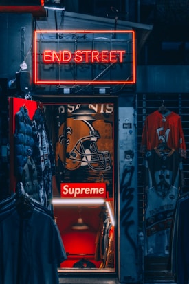 A dimly lit store entrance with a neon sign reading 'END STREET' above it. Inside, there are several items of clothing on display including jackets and jerseys. The store interior has a prominent 'Supreme' logo along with a large football helmet graphic on the wall. There are other visible details like hanging lights and graffiti on the door frame.