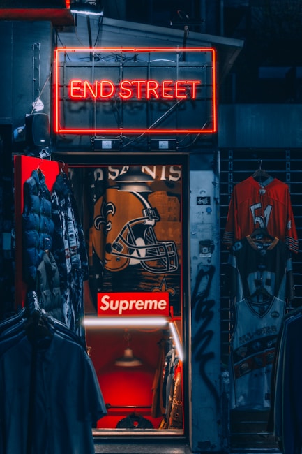A dimly lit store entrance with a neon sign reading 'END STREET' above it. Inside, there are several items of clothing on display including jackets and jerseys. The store interior has a prominent 'Supreme' logo along with a large football helmet graphic on the wall. There are other visible details like hanging lights and graffiti on the door frame.