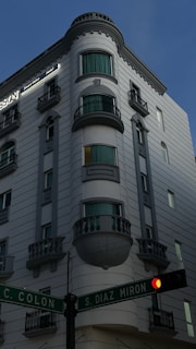 A multi-story building with a classic architectural style featuring rounded balconies and ornate railings. The facade is painted in light gray tones with windows covered by teal curtains. A street intersection is visible in the foreground, with signs indicating C. Colon and S. Diaz Miron. A traffic light is present and showing a red light.