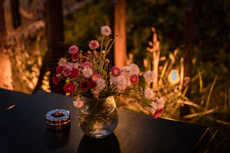 A close-up photo of an elegant metal flower vase with fresh blooms inside, sitting on a wooden table near a sunlit window.