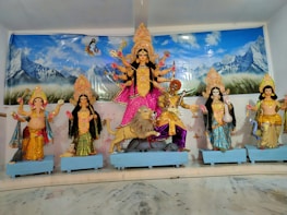 A vibrant display of Hindu deities, including a central figure of Goddess Durga, depicted with multiple arms and standing on a lion. Surrounding her are other deities with intricate traditional attire and ornaments, all set against a backdrop of mountains and a serene blue sky.