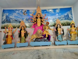 A vibrant display of Hindu deities, including a central figure of Goddess Durga, depicted with multiple arms and standing on a lion. Surrounding her are other deities with intricate traditional attire and ornaments, all set against a backdrop of mountains and a serene blue sky.