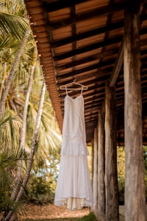 A white, lacy dress is hanging on a wooden hanger from a rustic roof, surrounded by green palm trees and other tropical foliage. The scene exudes a natural, serene atmosphere.