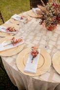 A round table is elegantly set with gold chargers, white napkins adorned with small floral arrangements, and clear wine glasses. In the center, a floral centerpiece combines dried grains and red berries in a terracotta vase. The tablecloth features a subtle checked pattern, and the setting is outdoors with visible grass and wicker chairs in the background.