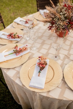 A round table is elegantly set with gold chargers, white napkins adorned with small floral arrangements, and clear wine glasses. In the center, a floral centerpiece combines dried grains and red berries in a terracotta vase. The tablecloth features a subtle checked pattern, and the setting is outdoors with visible grass and wicker chairs in the background.