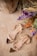 A pair of elegant, floral-embellished transparent sandals are placed on a textured beige surface. The sandals feature intricate golden and clear detailing. To the side, a vibrant arrangement of flowers in purple, orange, and green adds a touch of color.