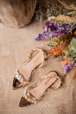 A pair of elegant, floral-embellished transparent sandals are placed on a textured beige surface. The sandals feature intricate golden and clear detailing. To the side, a vibrant arrangement of flowers in purple, orange, and green adds a touch of color.