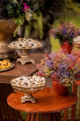 Several ornate, vintage-style dishes are arranged on a wooden table, filled with small desserts garnished with delicate leaves. To the side, a vibrant floral arrangement in a terracotta pot adds a burst of color with purple and red hues.