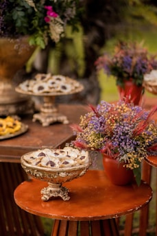 Several ornate, vintage-style dishes are arranged on a wooden table, filled with small desserts garnished with delicate leaves. To the side, a vibrant floral arrangement in a terracotta pot adds a burst of color with purple and red hues.