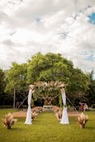 A lush green outdoor setting with a decorative wedding arch adorned with leaves and pampas grass. The arch is draped with white fabric, positioned in front of rows of wooden benches. The setting is surrounded by trees, under a partly cloudy sky, creating a serene and natural atmosphere.