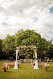 A lush green outdoor setting with a decorative wedding arch adorned with leaves and pampas grass. The arch is draped with white fabric, positioned in front of rows of wooden benches. The setting is surrounded by trees, under a partly cloudy sky, creating a serene and natural atmosphere.