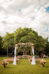 A lush green outdoor setting with a decorative wedding arch adorned with leaves and pampas grass. The arch is draped with white fabric, positioned in front of rows of wooden benches. The setting is surrounded by trees, under a partly cloudy sky, creating a serene and natural atmosphere.