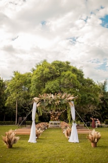A lush green outdoor setting with a decorative wedding arch adorned with leaves and pampas grass. The arch is draped with white fabric, positioned in front of rows of wooden benches. The setting is surrounded by trees, under a partly cloudy sky, creating a serene and natural atmosphere.