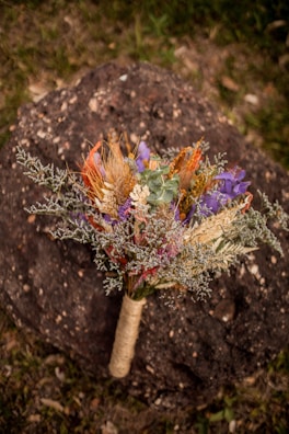 A vibrant bouquet featuring a mix of dried flowers and grasses placed on a textured rock surface. The bouquet includes hues of green, purple, orange, and red, wrapped with twine at the base. The backdrop is a natural setting with grass peeking through.