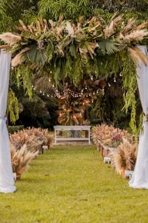An outdoor setting features an aisle lined with ornamental grasses and floral arrangements leading to an altar. A canopy adorned with greenery and pampas grass is suspended above, and string lights add a soft, glowing ambiance. The backdrop is lush with trees and foliage.
