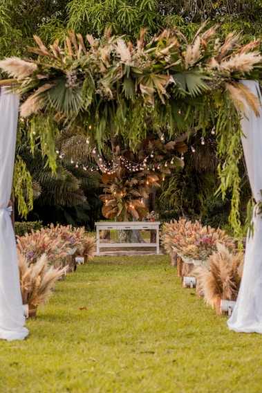 An outdoor setting features an aisle lined with ornamental grasses and floral arrangements leading to an altar. A canopy adorned with greenery and pampas grass is suspended above, and string lights add a soft, glowing ambiance. The backdrop is lush with trees and foliage.