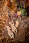 A pair of elegant, transparent sandals with floral embellishments is placed on a rustic wooden surface. Surrounding the sandals are dried flowers and a spool of twine, creating an earthy and vintage vibe.