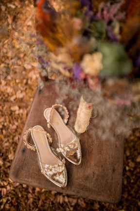 A pair of elegant, transparent sandals with floral embellishments is placed on a rustic wooden surface. Surrounding the sandals are dried flowers and a spool of twine, creating an earthy and vintage vibe.