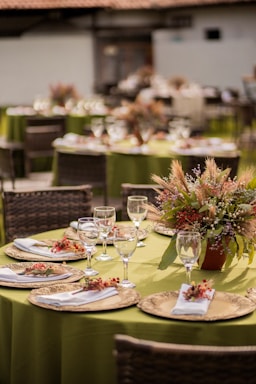 An elegantly set table features green tablecloths adorned with decorative plates, white napkins, and small floral arrangements. The setting includes wine glasses and a centerpiece made of dried flowers and foliage, creating a rustic yet sophisticated ambiance.