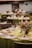 An elegantly set table features green tablecloths adorned with decorative plates, white napkins, and small floral arrangements. The setting includes wine glasses and a centerpiece made of dried flowers and foliage, creating a rustic yet sophisticated ambiance.