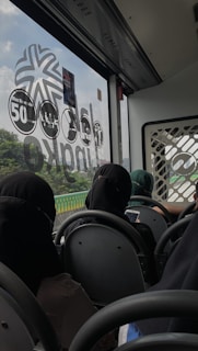 The interior of a bus is visible with passengers seated in rows. The bus window showcases a view of greenery outside and has various symbols and numbers on it. The passengers, wearing various clothing, are engaged on their mobile devices or looking out the window.