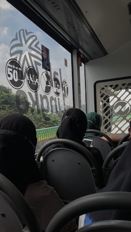 The interior of a bus is visible with passengers seated in rows. The bus window showcases a view of greenery outside and has various symbols and numbers on it. The passengers, wearing various clothing, are engaged on their mobile devices or looking out the window.