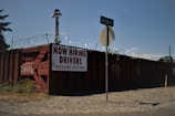 A barbed wire fence surrounds a perimeter with a large, rusty red shipping container that has graffiti on it. A sign on the container reads 'NOW HIRING DRIVERS INQUIRE WITHIN,' and a street sign for 'Tule Lake Rd S' is visible in front. The ground is covered with gravel and there is some overgrown grass.