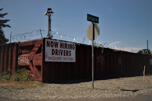 A barbed wire fence surrounds a perimeter with a large, rusty red shipping container that has graffiti on it. A sign on the container reads 'NOW HIRING DRIVERS INQUIRE WITHIN,' and a street sign for 'Tule Lake Rd S' is visible in front. The ground is covered with gravel and there is some overgrown grass.