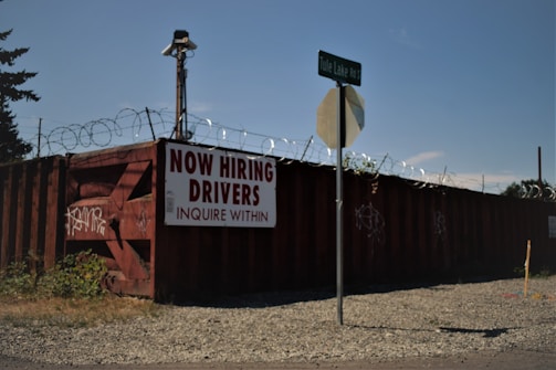 A barbed wire fence surrounds a perimeter with a large, rusty red shipping container that has graffiti on it. A sign on the container reads 'NOW HIRING DRIVERS INQUIRE WITHIN,' and a street sign for 'Tule Lake Rd S' is visible in front. The ground is covered with gravel and there is some overgrown grass.